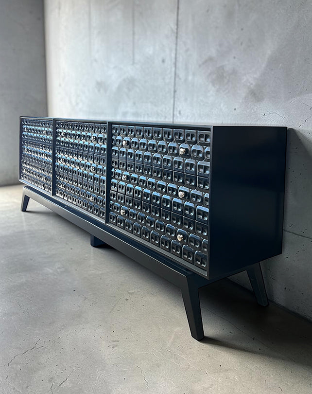 Modern black console table with metallic door panels against a concrete wall.
