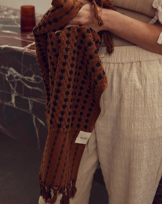 Person holding a brown knitted blanket with a brand label against a blurred background