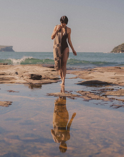 Woman walking on a rocky beach with a towel, reflected in the water.