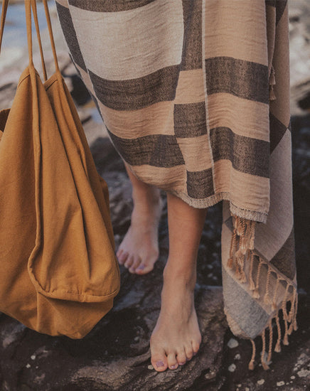 Person wrapped in a plaid towel on a rocky beach with a mustard yellow bag.