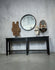 A black wooden console table against a textured gray concrete wall, styled with a round black-framed mirror, a carved wooden sculpture, a white hat on a stand, and a decorative mask.
