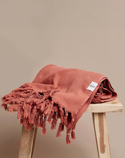 Reddish-brown blanket with tassels on a wooden stool against a beige background