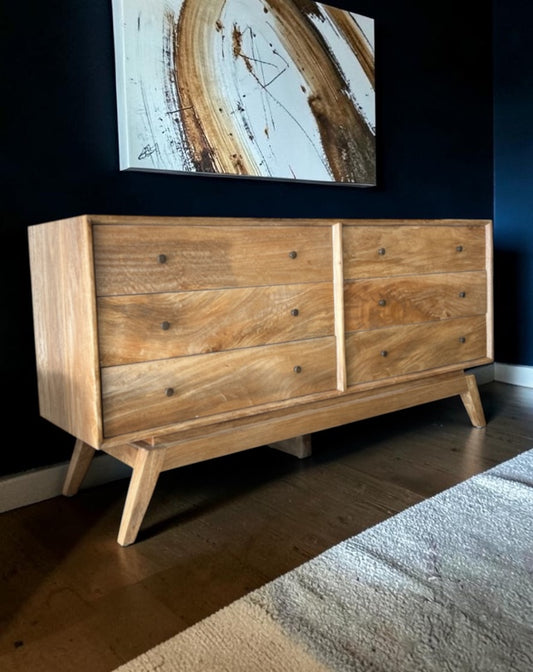 A mid-century modern wooden sideboard with a natural oak finish, featuring three drawers on the left and two larger drawers on the right, supported by angled legs, set against a dark navy wall in a styled room.