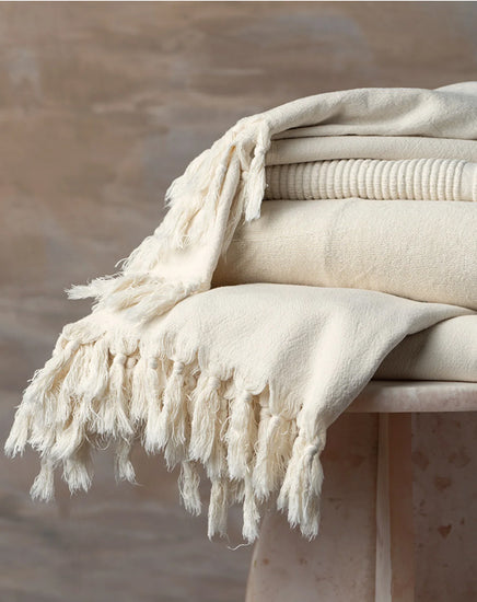 Beige blanket with tassels draped over a surface against a neutral background
